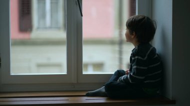 Apartman penceresinin yanında oturan çocuk evden yağmur manzarasına bakıyor. Kapalı küçük bir çocuk soğuk bir günde süveter giyer.