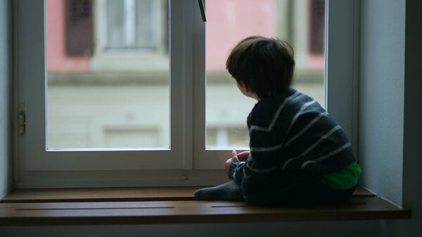 Child sitting by apartment window staring at drizzle view from home. Confined little boy contemplating view during cold day, wearing sweater