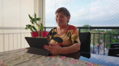 Evde tablet kullanan mutlu yaşlı kadın. Yaşlı olgun bir kadın apartman balkonundaki modern teknoloji ekranına bakıyor.
