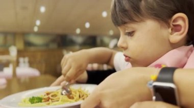 Küçük çocuk lokantada peçete önlüğüyle spagetti yerken yetişkinlerden yardım alıyor.