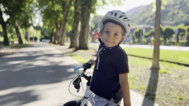 若い男の子は バックグラウンドの車や山と一緒にアベニューの木陰で日当たりの良い日に彼の自転車の写真のためのヘルメットポーズを着用 — ストック動画