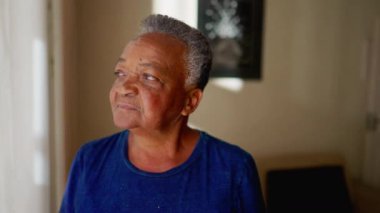 Düşünceli yaşlı kadın pencerenin kenarında dikilip eve bakıyor. 70 'lerdeki Afrikalı Amerikalı yaşlı bir insanın düşünceli ifadesi.