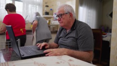 Ev mutfağı ortamında modern teknolojiyi kullanan dizüstü bilgisayarın önündeki yaşlı adam. Beyaz erkek, üst düzey, evde bilgisayarla meşgul.