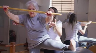 Pilates hocası, fizyoterapi sınıfındaki yaşlı bir kadına tahta sopa ve esneme bacağıyla rehberlik ediyor.