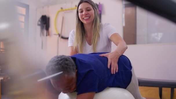 普拉提指导员帮助一名巴西老年黑人妇女在理疗室内进行球类运动 — 图库视频影像