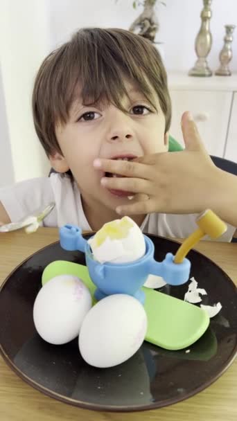 陽気な少年は 彼のお気に入りの子供をテーマにした卵ホルダーに置かれた沸騰した卵を味わうためにスプーンを使用し 食事の時間を楽しいものにします — ストック動画