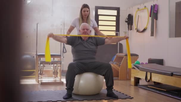 Senior Man Exercising Elastic Bands Seated Pilates Ball Being Oriented — Stock Video