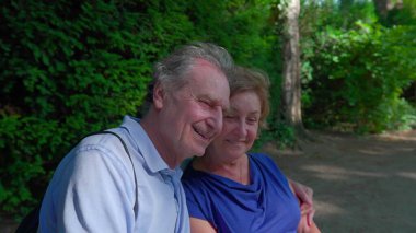 Parktaki bankta güneşli günün tadını çıkaran neşeli yaşlı çift. Koca yaşlı karısına sarıldı. Gülümsüyor ve dışarıda gülüyor. Mutlu bir evlilik.