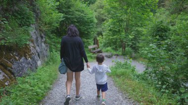 Anne ve çocuğun arkası, hafta sonu aktiviteleri sırasında doğanın yolunda el ele yürüyorlar. Anne ve küçük çocuk yürüyüş gününün tadını çıkarıyor.