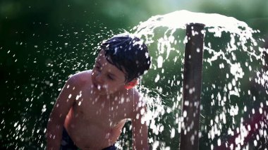 Çocuk, halka açık havuz suyu çeşmesinde yavaş çekimde 120fps ile kendini yeniliyor. Küçük çocuk sıcak yaz gününde su damlacıklarının keyfini çıkarıyor.