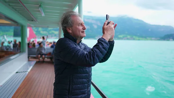 Senior man traveling by boat pulling out cellphone to take a photo of lake and mountain view. Older man retired man exploring new horizons filming nature with smartphone device
