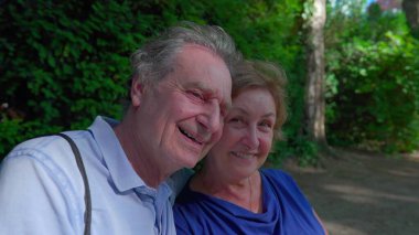 Parktaki bankta güneşli günün tadını çıkaran neşeli yaşlı çift. Koca yaşlı karısına sarıldı. Gülümsüyor ve dışarıda gülüyor. Mutlu bir evlilik.