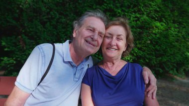 Mutlu yaşlı çift güneşli yaz gününde parkta bankta oturuyor. Yaşlı kocanın kollarına sarılıp poz vermesi, yakın plan yüzler, yanak yanağa.