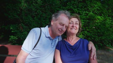 Mutlu yaşlı çift güneşli yaz gününde parkta bankta oturuyor. Yaşlı kocanın kollarına sarılıp poz vermesi, yakın plan yüzler, yanak yanağa.