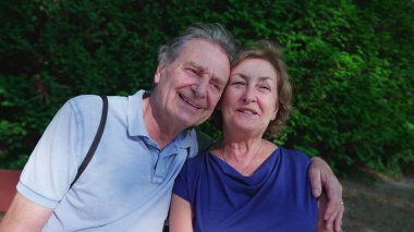 Mutlu yaşlı çift güneşli yaz gününde parkta bankta oturuyor. Yaşlı kocanın kollarına sarılıp poz vermesi, yakın plan yüzler, yanak yanağa.