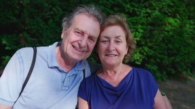 Mutlu yaşlı çift güneşli yaz gününde parkta bankta oturuyor. Yaşlı kocanın kollarına sarılıp poz vermesi, yakın plan yüzler, yanak yanağa.