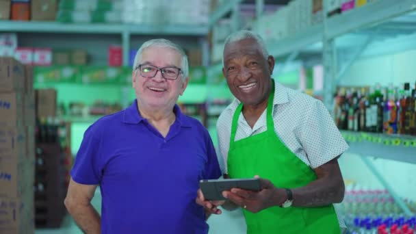 各色人等的巴西超市连锁店资深员工带着平板电脑和制服对着摄像机微笑 非裔美国人老年雇员和一个高加索人一起笑 — 图库视频影像