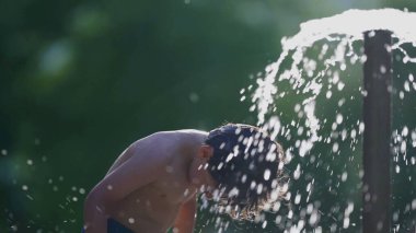 Küçük çocuk, yavaş çekim 120 fps yüzme havuzu çeşmesinde kendini yeniliyor. Sıcak yaz gününde su damlacıklarının keyfini çıkaran bir çocuk.