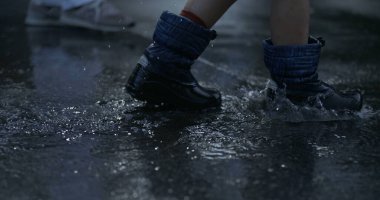 Yakın plan yağmur botları su birikintilerinde yürüyor her yere damlacıklar sıçrıyor yağmurlu bir öğleden sonra 800 fp hızla