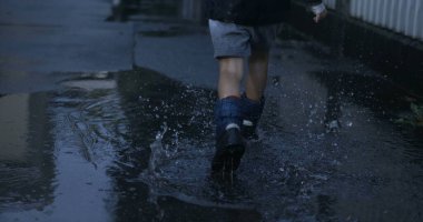 Çocuk sevinçle yağmur botlarıyla su birikintisine koşuyor.