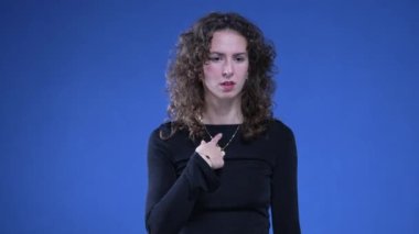 İnançsızlık ve şaşkınlık içinde kendini işaret eden bir kadın mavi arka planda duruyor. 20 'li yaşlarda biri suçlamalara tepki veriyor