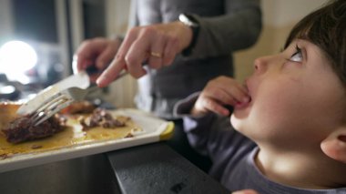 Anne yemek hazırlarken küçük çocuk da mutfakta onu izliyor. Otantik gerçek bir aile hayatı sahnesi, o yemek pişirirken ebeveynin oğluna yemek verdiği sahne.