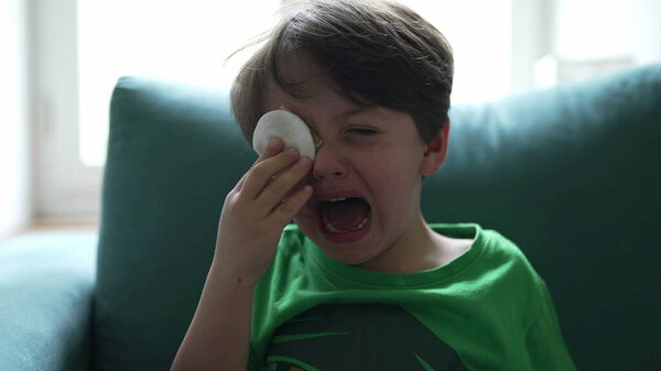 Crying small boy with hurt eye emergency, open mouth tearful child feeling pain after accident, hand covering eye with cotton