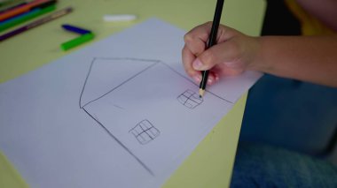 Yakın plan çocuk eli, geleneksel bir ev, iki pencere ve kağıt üzerinde kurşun kalemle ön kapı çiziyor, yaratıcılık oyun zamanı çocukluk kavramı