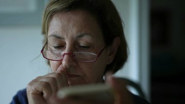 Yaşlı kadın akıllı telefon cihazıyla internetten içeriği okurken burnunu ve yüzünü kaşır. Konsantre Kafkasyalı kişi