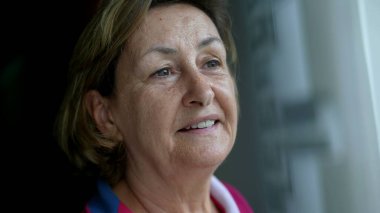 Yakın plan mutlu, düşünceli yaşlı kadın pencerenin kenarında durmuş manzaraya bakıyor ve sessiz bir sevinç hissediyor. Emekli Kafkasyalı yaşlı bayanın düşünceli ifadesi.