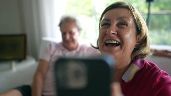 Happy grandmother laughing and smiling while interacting with relatives in video call. Long-distance communication between grandparents seated on couch speaking with family
