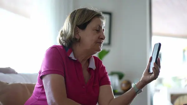 stock image Elderly Woman Greeting Family on Video Call from Sofa, Joyful Grandmother Video Chatting with Relatives at Home waving hello and sending kisses