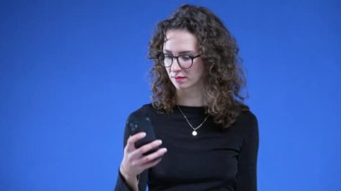 Cep telefonu çeken bir kadın mavi arka planda dururken mesaj alıyor. Okuma gözlüğü takan ve elinde telefon tutan biri.