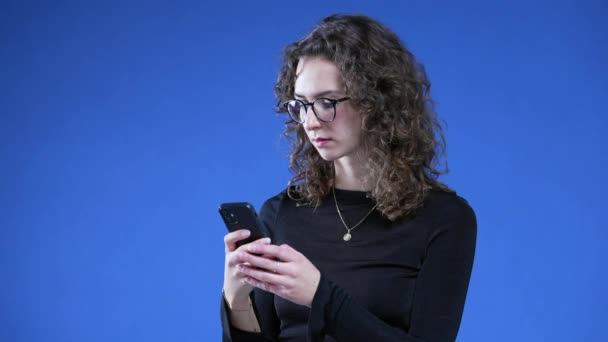 站在蓝色背景下使用手机的女人 20多岁的女性戴眼镜 带着中性表情在电话中发短信 — 图库视频影像