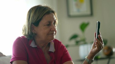 Evdeki kanepede otururken ekranda akıllı telefon cihazını tutan son sınıf öğrencisi kadın, hoparlör dinleyen Kafkasyalı yaşlı kadının nötr ifadesi.