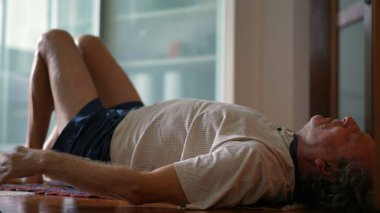 Sabahları kıdemli bir adam bacaklarını açıyor. Yaşlı, emekli, beyaz, sırt üstü uzanmış egzersiz yapıyor ve vücuduna bakıyor.