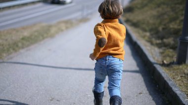 Back of excited small boy sprinting outside during autumn day, Child wearing yellow pullover, jeans, and boots runs clipart