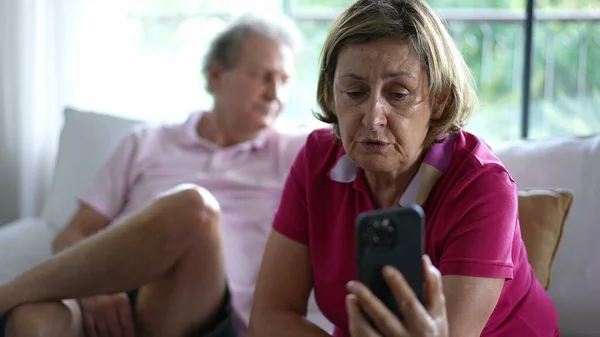 Senior grandmother speaking with family relatives on video communication, holding smartphone device seated at home couch sofa next to her husband