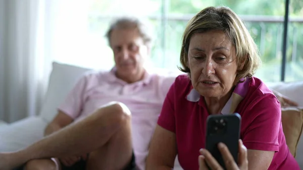 Senior grandmother speaking with family relatives on video communication, holding smartphone device seated at home couch sofa next to her husband