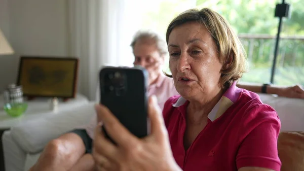 Senior woman holding smartphone device speaking with family member using video communication to speak in long-distance. Grandparents speaking with relatives