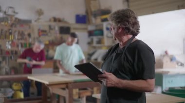Küçük işletme sahibi marangoz dükkânı elinde tablet, yerel marangoz dükkanının online siparişlerini tarıyor, modern teknoloji kullanarak marangoz önlüğü giyiyor.