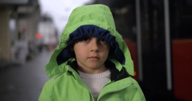 Yağmur altında kaldırımda dikilen yeşil yağmurluk giymiş ve kameraya tarafsız bir ifadeyle bakan küçük bir çocuğun portresi. Yağmur mevsiminde çocuk yüzü