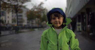 Yağmur damlacıkları süper yavaş çekimde düşerken yeşil yağmurluk giymiş çocuk arka planda şehir sokağında dikiliyor kameraya gülümsüyor.