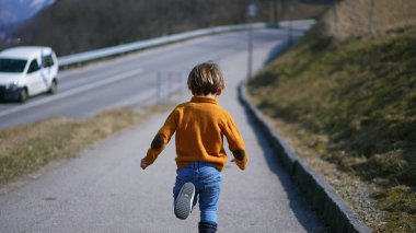 Arkadan, sarı kazaklı, kot pantolonlu ve botlu neşeli bir çocuk sonbahar manzarası boyunca koşuyor. Canlı koşu, gevrek sonbahar havasının heyecanını somutlaştırır.