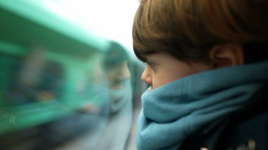 Trenin camına yaslanan küçük çocuk hızla geçen manzaraya bakıyor. Eşarp takmış çocuk hareketli manzaraya bakıyor