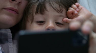 Yakın plan çocuk annesiyle telefon ekranına bakıyor. Çevrimiçi içerik tüketen otantik çocuk