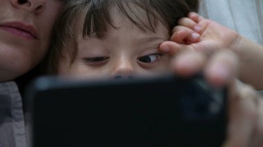 Yakın plan çocuk annesiyle telefon ekranına bakıyor. Çevrimiçi içerik tüketen otantik çocuk