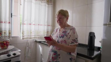 Mutfak lavabosuna yaslanırken cep telefonu kullanan yaşlı bir kadın. 80 'lerde İnternet' te gezinen yaşlıların ev içi yaşam tarzı