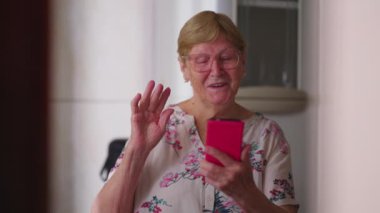 Evdeki video konferansında konuşan aile üyesine el sallayan kıdemli bir kadın. Akıllı telefon aygıtı ekranına bakarak akrabalarla konuşan otantik yaşlı kadın