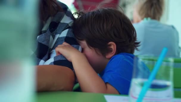 疲惫的小男孩靠在妈妈的胳膊上 在餐馆等着食物的到来 感到厌烦 孩子粘在父母身上 揉着脸 — 图库视频影像